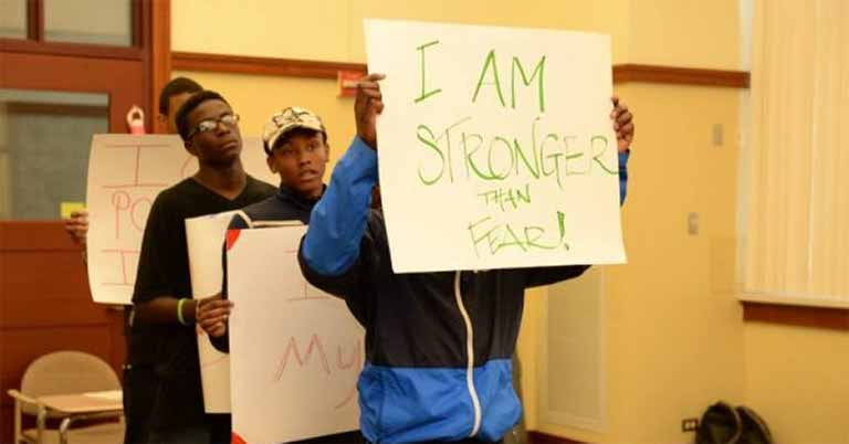 Four young people stand in a line with signs in hand. The front sign reads "I am stronger than fear!"