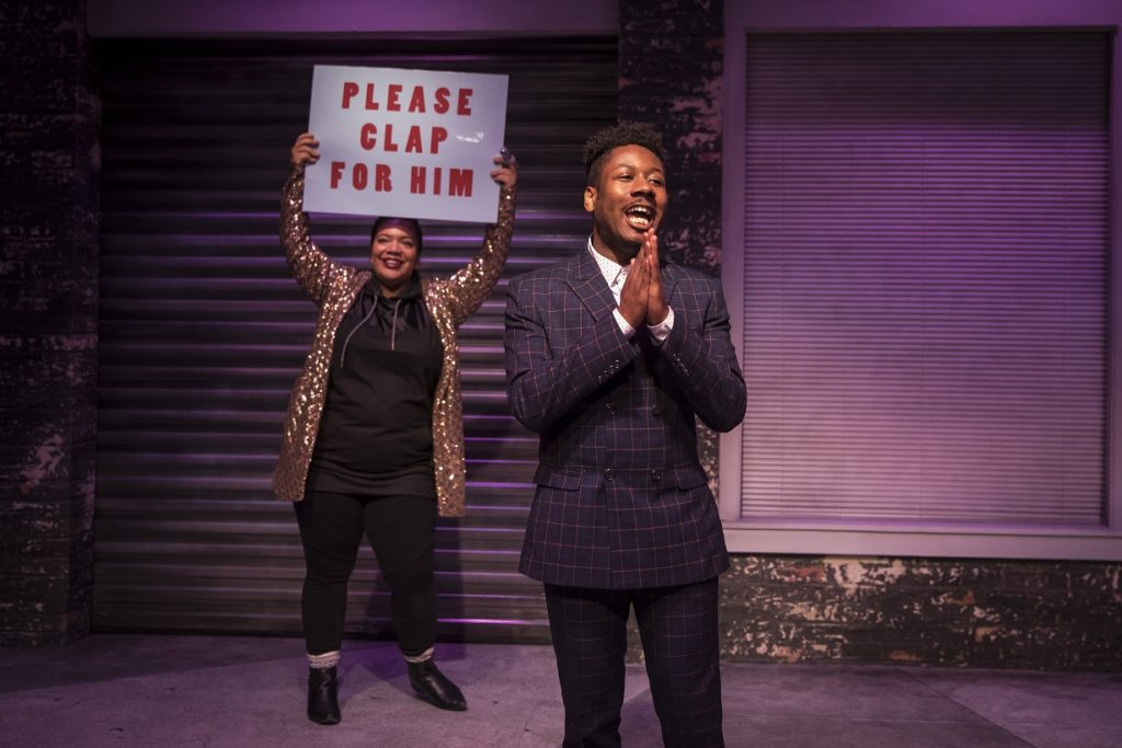 Jerome Beck standing with Sasha Smith in the background with a sign that reads, "Please clap for him."